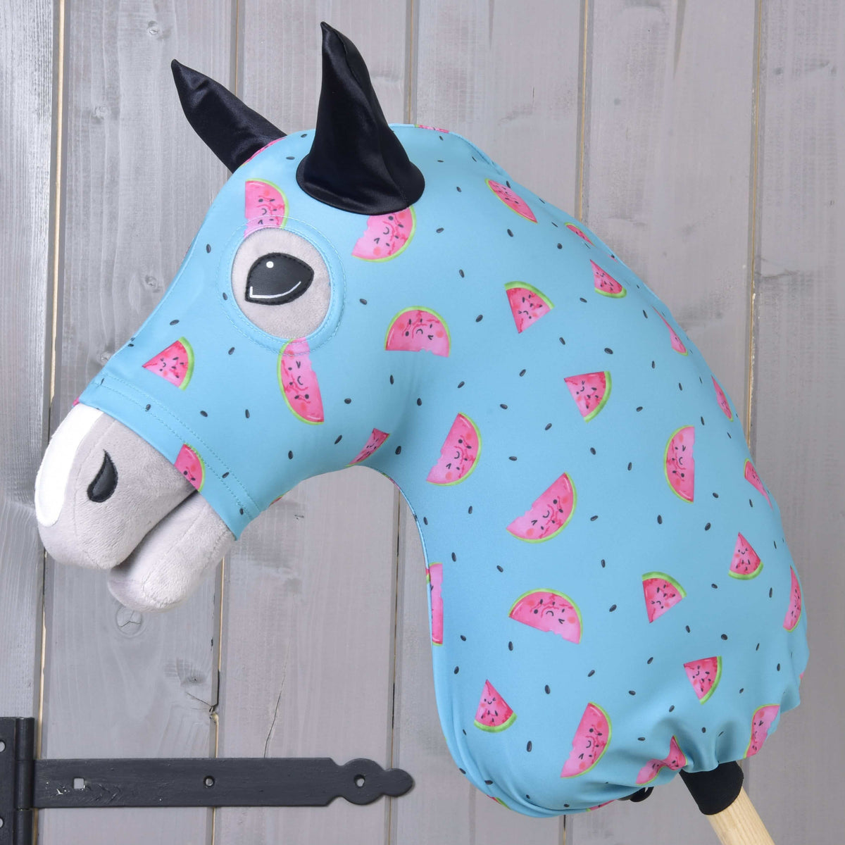 The Hobby Horse LarDen protective suit Melon (Lycra) features a washable blue cover with pink watermelon slices and black seeds, designed for plush hobby horse heads. Ideal for protection, shown on a wooden stick against a light wood paneled wall.
