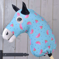 The Hobby Horse LarDen protective suit Melon (Lycra) features a washable blue cover with pink watermelon slices and black seeds, designed for plush hobby horse heads. Ideal for protection, shown on a wooden stick against a light wood paneled wall.