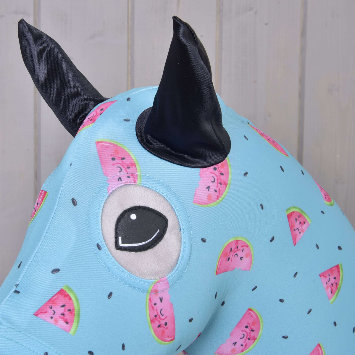 A close-up of the Hobby Horse LarDen protective suit Melon (Lycra), a light blue washable lycra cover with watermelon patterns, one large embroidered eye, and two black fabric ears, set against a light wooden background.