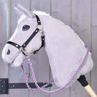 The Hobby Horse LarDen "Reins knitted pinkie blue" is a gray plush hobby horse with a black bridle and hand-braided pink, blue, and white reins on a wooden stick, shown against a light wooden background.