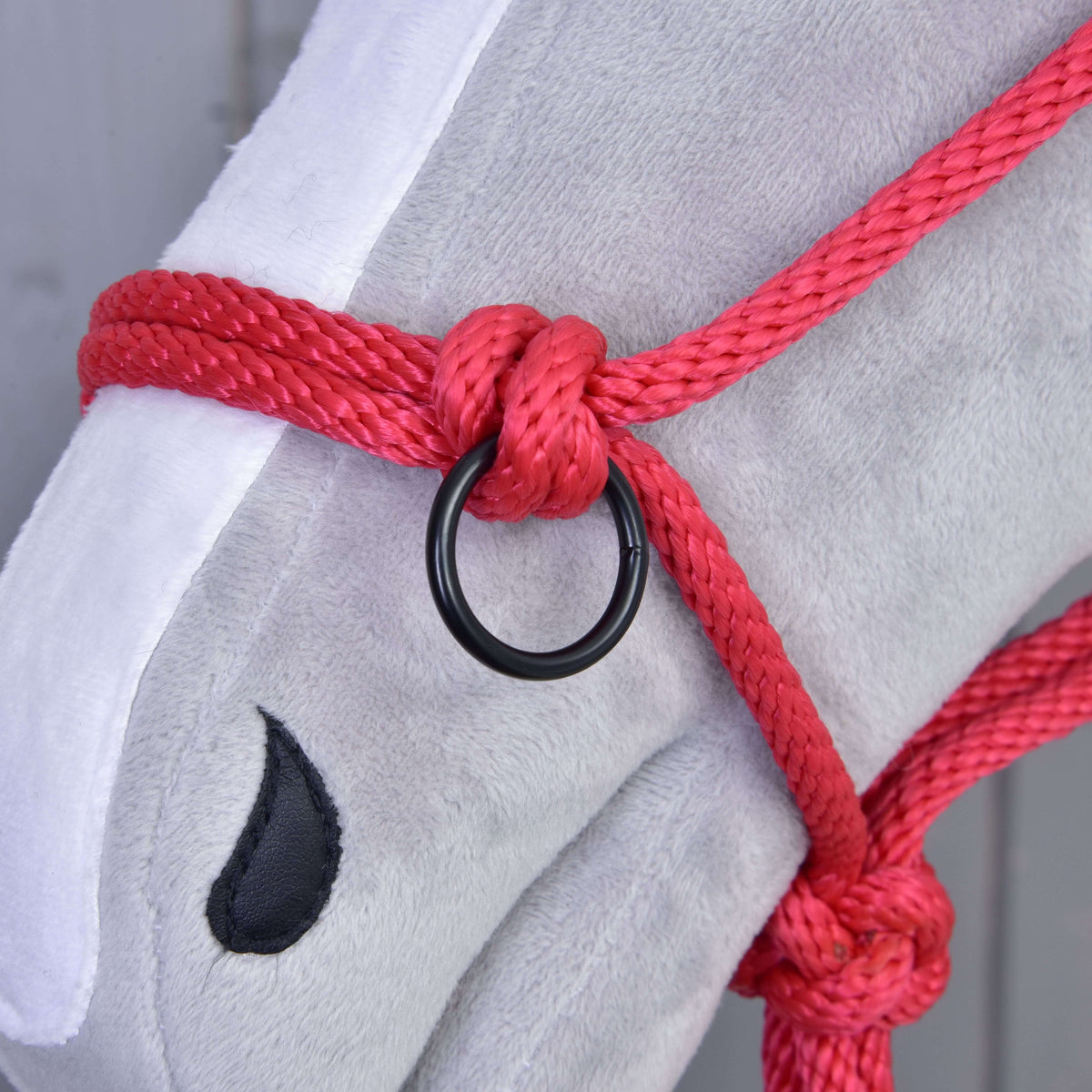 Close-up of a plush hobby horse head with a black stitched eye, wearing the Hobby Horse LarDen Rope halter red with a black metal ring. The background is blurred gray wood.