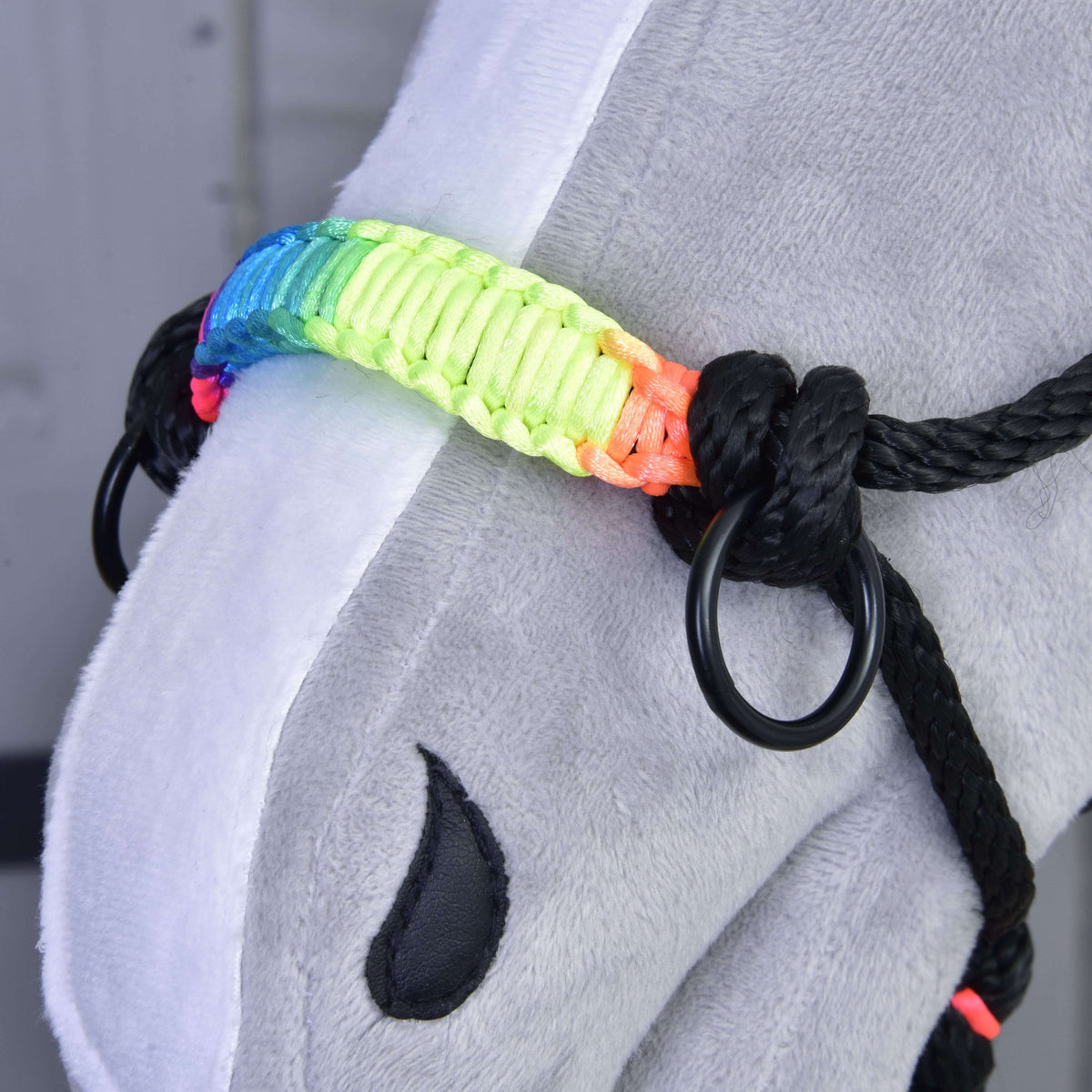 Close-up of a grey plush hobby horse by Hobby Horse LarDen, wearing the Rope halter Rainbow with metal rings and a vibrant rainbow noseband. A black teardrop-shaped marking is visible on its face.