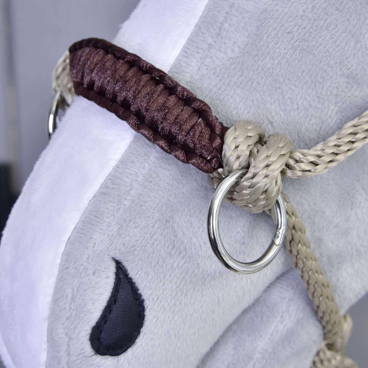 Close-up of a plush horse's face wearing the Rope halter Nature by Hobby Horse LarDen, featuring beige and brown braiding, a metal ring, and a black embroidered eye detail.