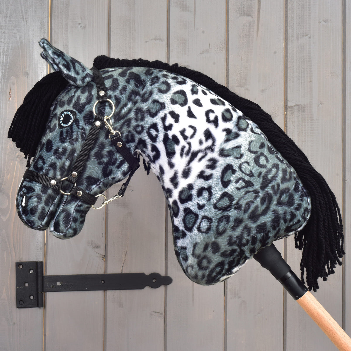The Hobby Horse LarDen "Hobby Horse Picasso with Black Halter" features a gray-and-black leopard-print plush coat, a black yarn mane, and a black halter. It is shown mounted on a wooden stick, set against a gray wooden wall with a black metal hinge.