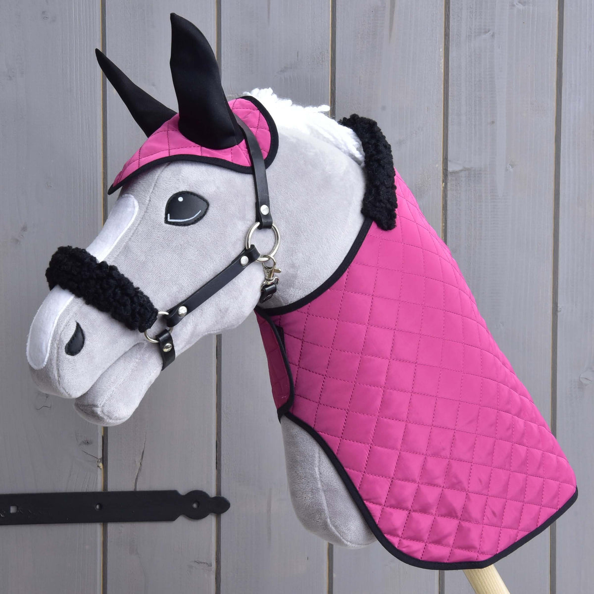 The Hobby Horse LarDen Set Pink Beauty features a plush toy hobby horse with a quilted pink blanket, matching ear bonnet, and a fur-trimmed halter—essential accessories—posing before wooden planks.