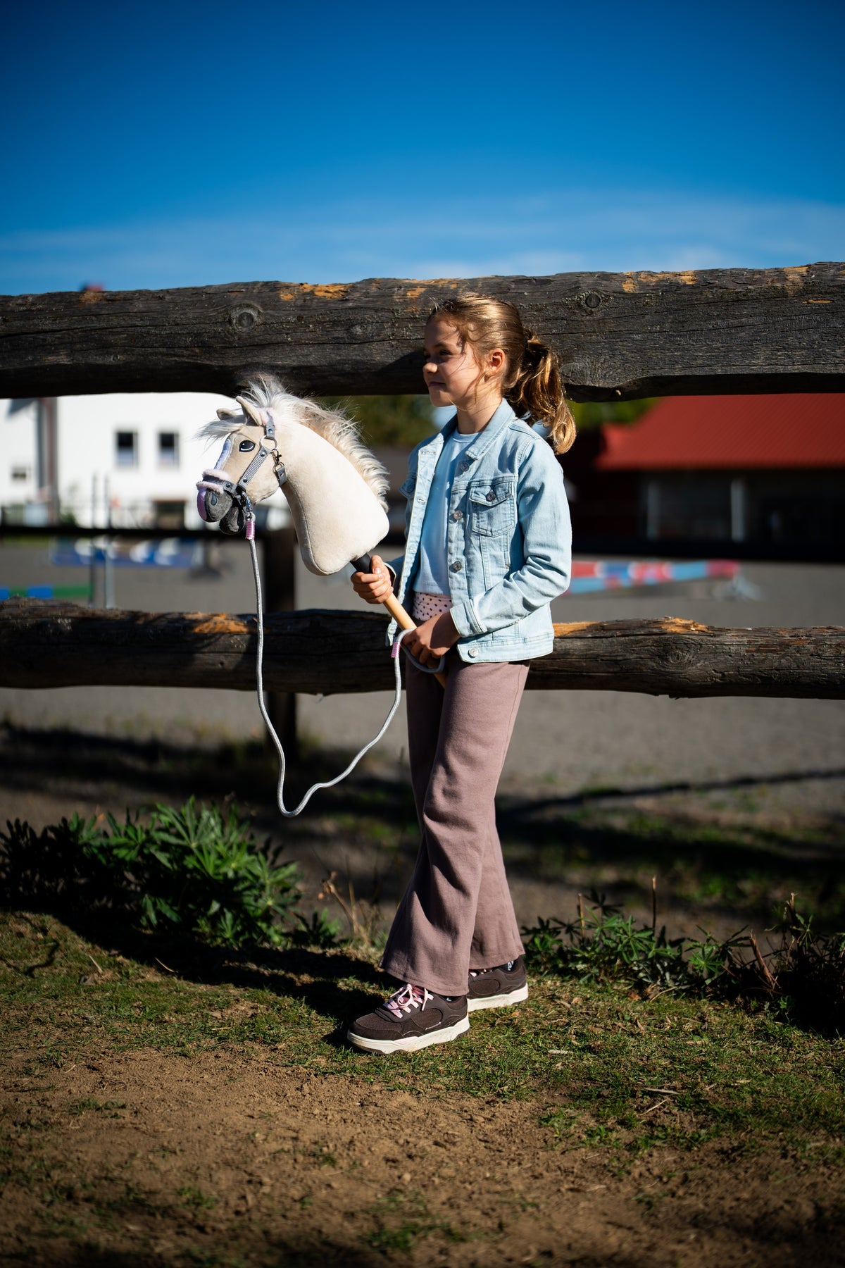 Lifestyle photo: a girl in a light denim jacket and mauve trousers stands at a wooden paddock fence holding the Hobby Horse Pixie with Black Halter by Hobby Horse LarDen - beige plush head with a white yarn mane - shown here with the Halter grey with pink fur and the rope stripe Tizian lead.