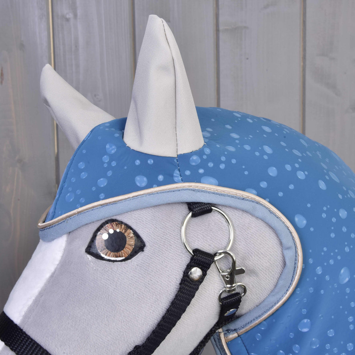 A close-up of a Hobby Horse LarDen Raincoat Dolly (softshell) head, featuring a blue water droplet-patterned raincoat and black halter, set against light wooden panels—ideal for all-weather play.