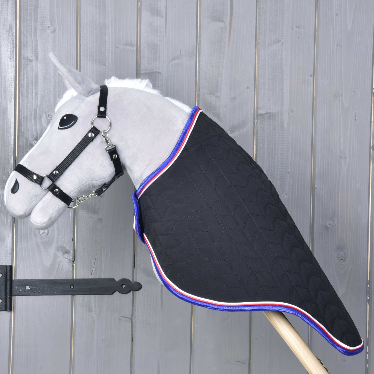 A gray Hobby Horse LarDen toy, featuring the Travel Rug Luxury—a black quilted blanket with red, white, and blue trim—stands on a wooden stick against a rustic wooden background.