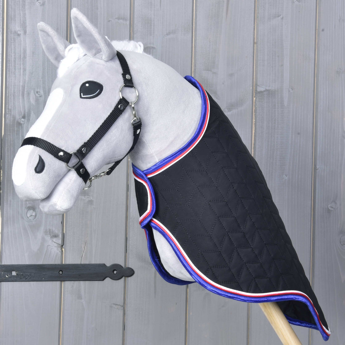A gray hobby horse toy with a black bridle features the Hobby Horse LarDen Travel Rug Luxury—quilted black, trimmed in red, white, and blue—posed against a wooden wall background.