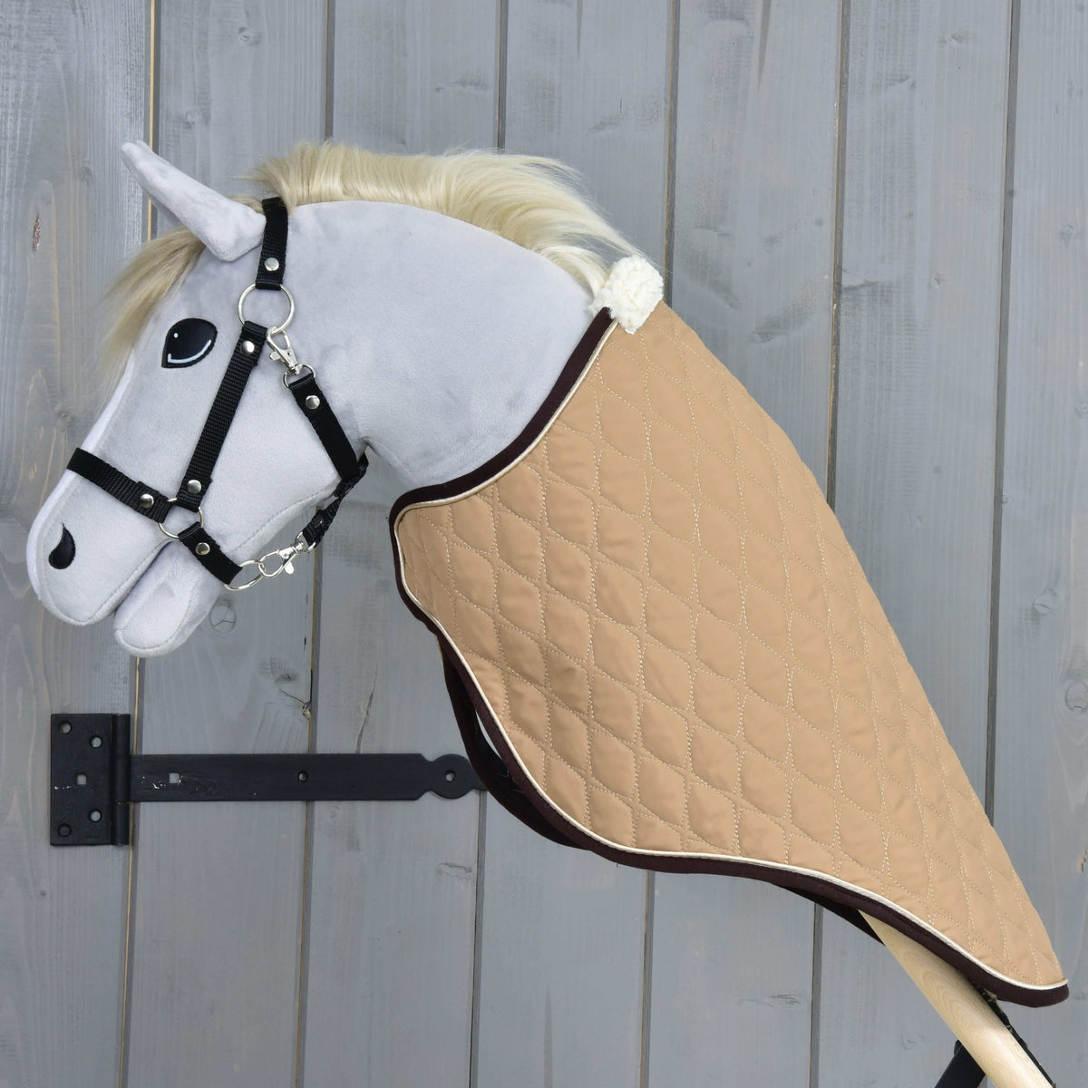 Det beige quiltede transportdækken set fra siden på en grå kæphest af plys, hvor det dækker halsen helt ned til kæppen