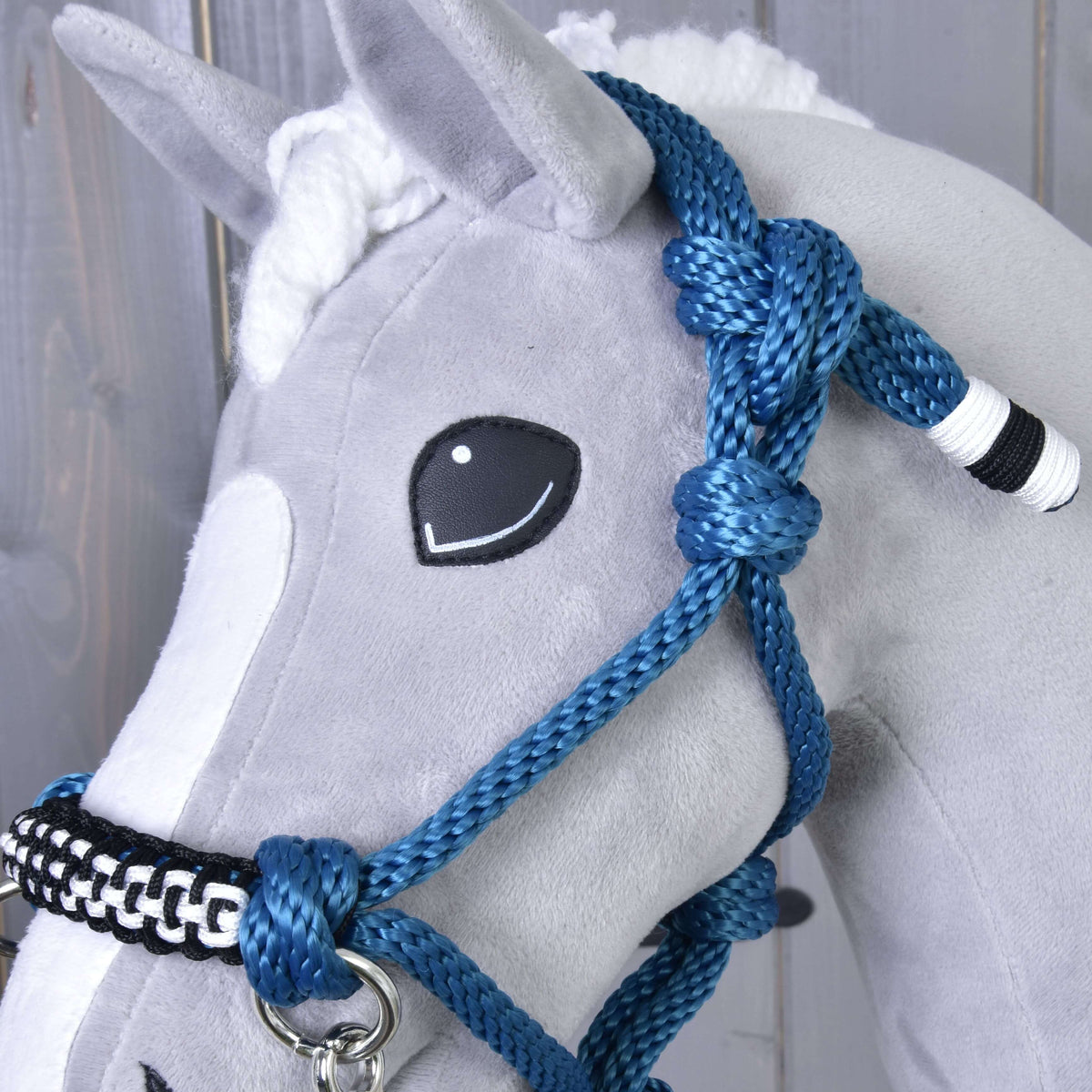 Close-up of a gray plush horse by Hobby Horse LarDen, featuring the Rope set Chess—a handmade halter, reins, cordeo, and blue braided rope with white and black accents. The horse has a stitched smile and white mane against a wooden background.