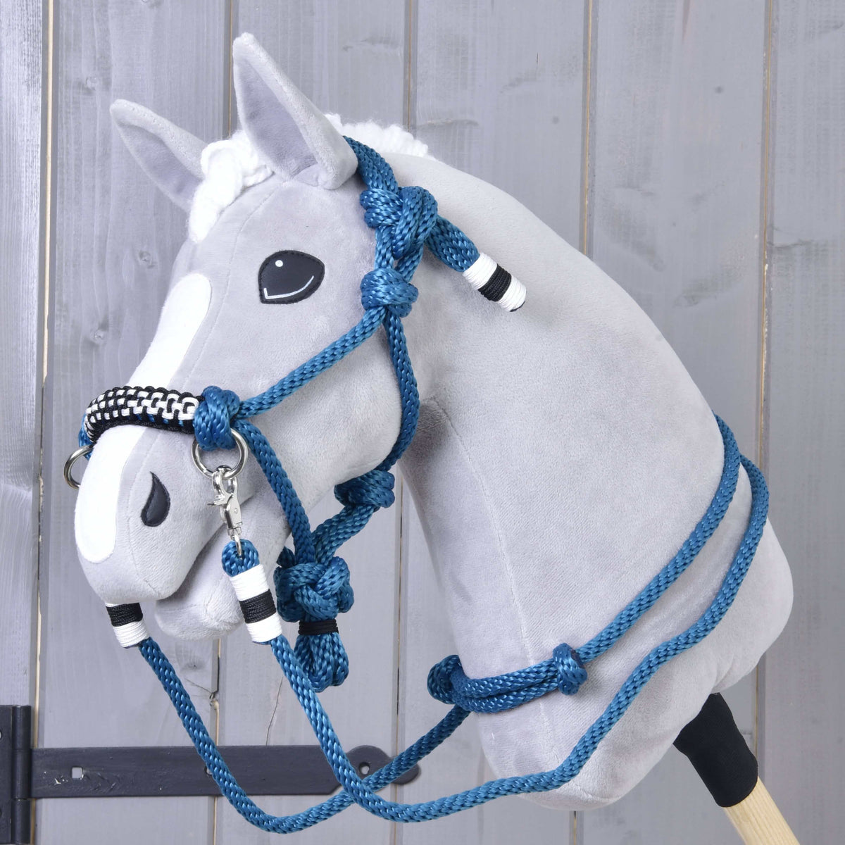 The Rope set Chess by Hobby Horse LarDen is a plush hobby horse with a gray face, white blaze, and blue halter reins cordeo with black and white accents, displayed against a wooden wall background.