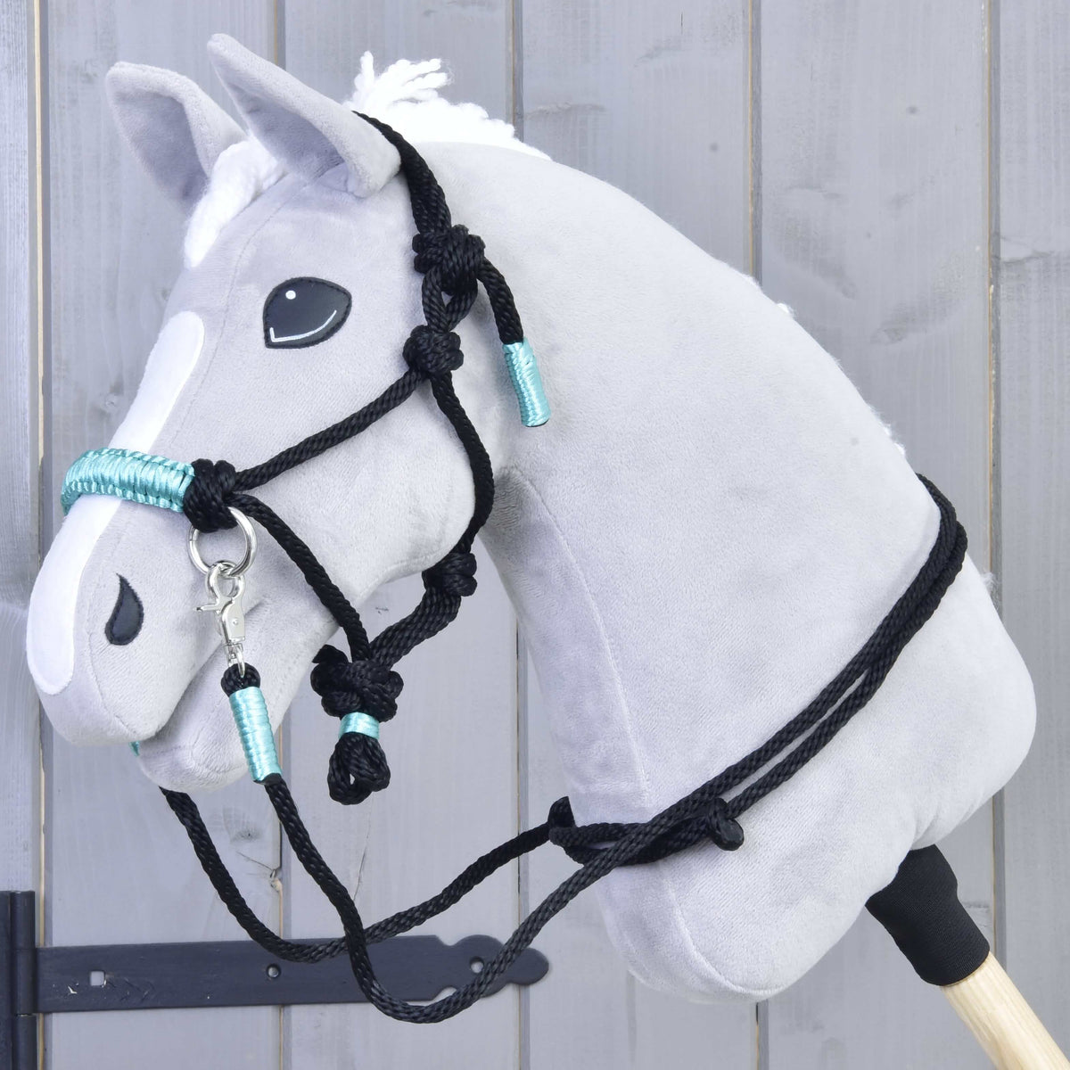 The Hobby Horse LarDen Rope set Turquoise is shown on a grey plush hobby horse with a white mane, black bridle, and turquoise accents mounted on a wooden stick in front of a gray wooden wall.