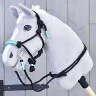 The Hobby Horse LarDen Rope set Turquoise is shown on a grey plush hobby horse with a white mane, black bridle, and turquoise accents mounted on a wooden stick in front of a gray wooden wall.