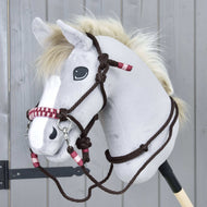 A gray hobby horse with a plush head, white mane, and dark eyes wears the Hobby Horse LarDen Rope set York bridle and halter with red and white accents, standing in front of a gray wooden background.