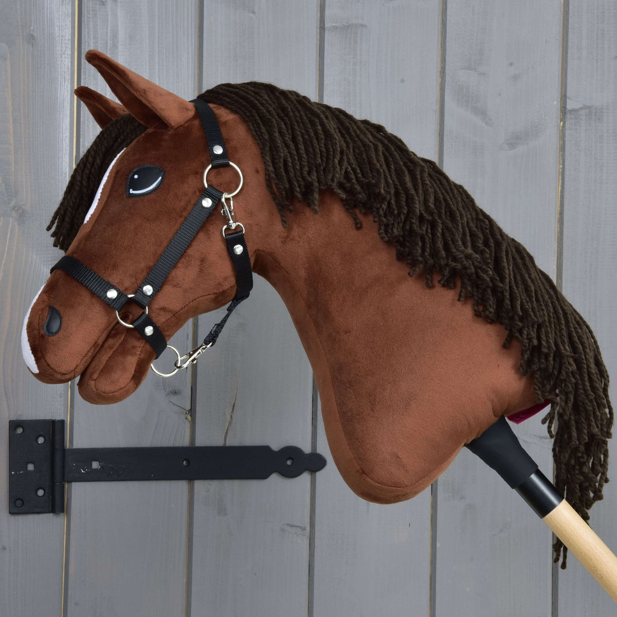 The Hobby Horse LarDen "Hobby Horse Rocky with Black Halter," a brown plush stick horse, is shown next to a gray wooden wall.
