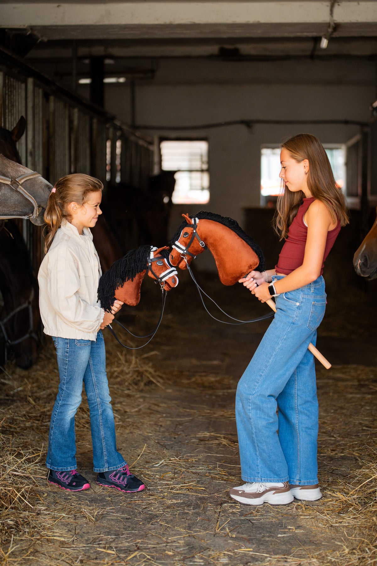 Lifestyle photo: two girls face each other in a stable aisle, each holding a Hobby Horse Rocky by Hobby Horse LarDen - chestnut brown plush head with a white blaze and a black yarn mane - shown here with the black complete bridle with fur and reins.