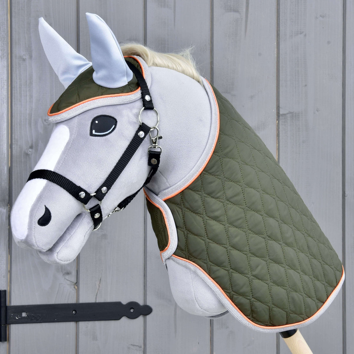 The Hobby Horse LarDen Set Moos features a gray hobby horse with black bridle, green quilted blanket, and light blue ear bonnet on a wooden stick, shown against a gray wooden wall.