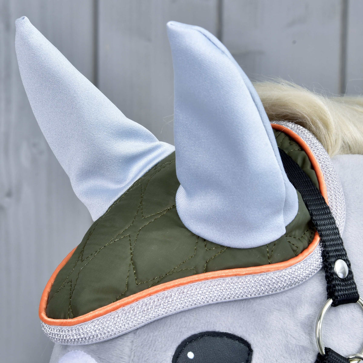Close-up of the Set Moos soft toy horse’s head by Hobby Horse LarDen, featuring an olive green and orange ear bonnet, two light gray pointed fabric ears, and a cute horse decoration against a light gray wooden background.