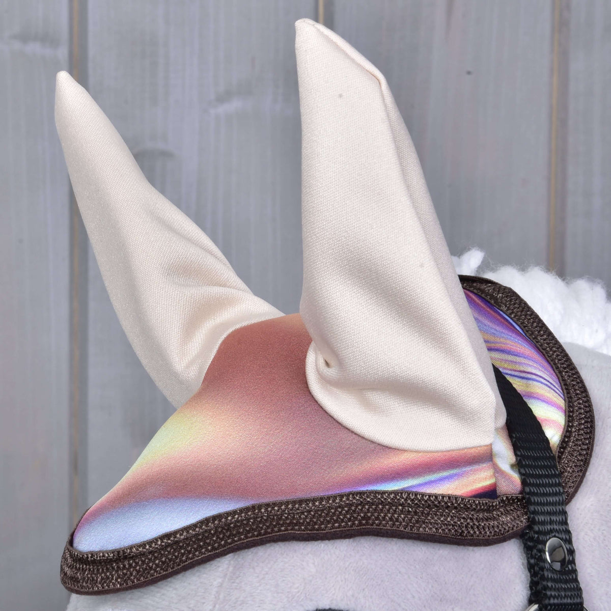 Close-up of Set Valery by Hobby Horse LarDen—a toy with shiny, iridescent ear bonnet and two white upright fabric ears. Blurred wooden panels in the background.