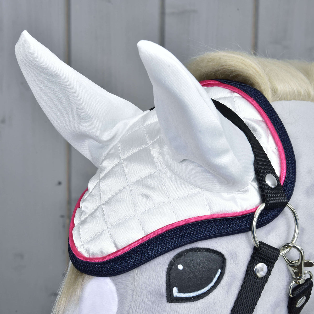 Close-up of Hobby Horse LarDen's Set Beautiful Star: a gray toy horse with white quilted ear bonnet featuring pink trim and navy border, black bridle straps, cartoon eyes, set against light wood paneling—perfect horse protection.