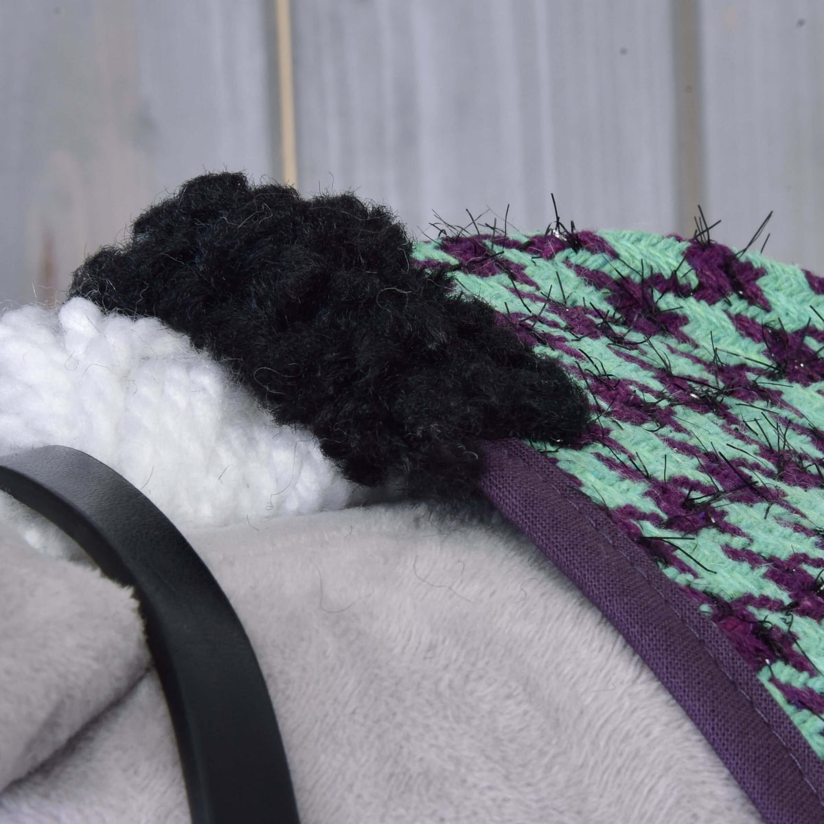 Close-up of Hobby Horse LarDen's Set Harmony Green head, showing a black and white yarn mane, green and purple textured fabric, gray plush surface, and black halter strap against a wooden background.