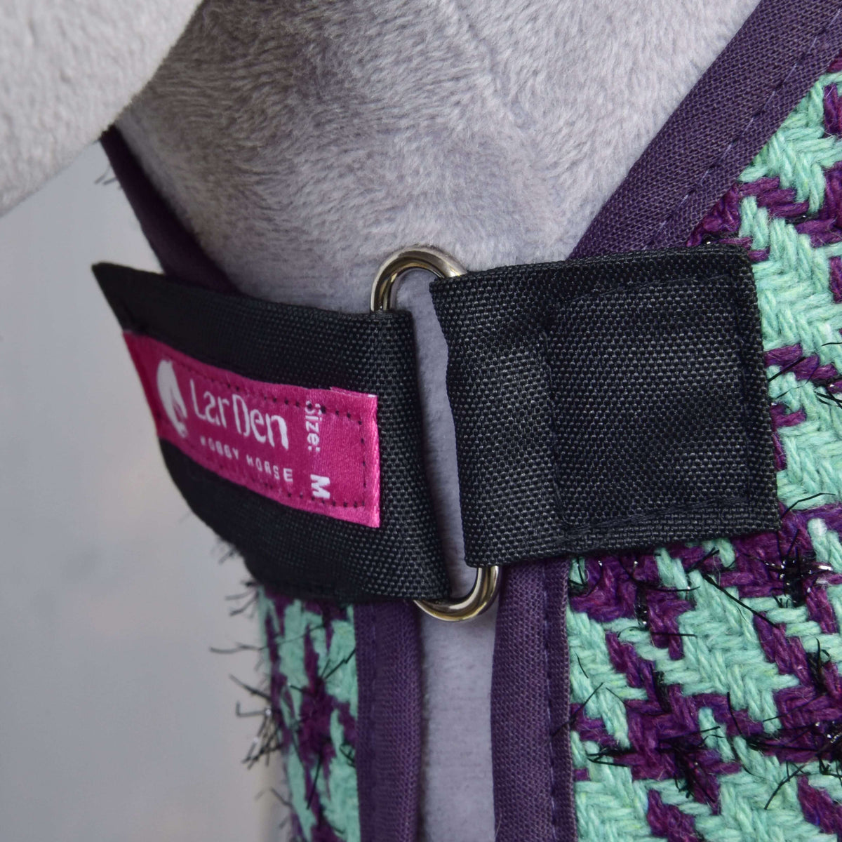 Close-up of a Hobby Horse LarDen Set Harmony Green harness in purple and green weave, black strap with metal D-ring, and pink M size tag. Worn by a light gray hobby horse.