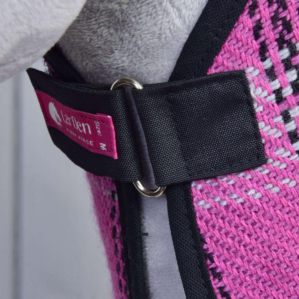 Close-up of Set Harmony Pink by Hobby Horse LarDen: pink woven fabric with a black halter strap, metal ring, and pink tag. Background hints at grey and black details from a garment or hobby horse accessory, possibly a stable blanket.