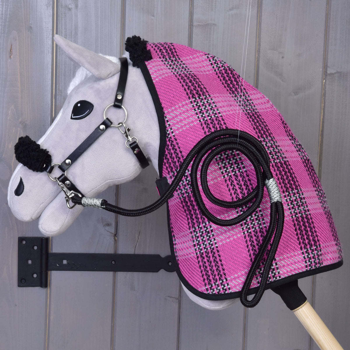 A Hobby Horse LarDen "Set Harmony Pink" stick toy in gray is mounted on a wooden wall, dressed in a pink and black plaid stable blanket with a black halter and matching coiled lead rope on its side.