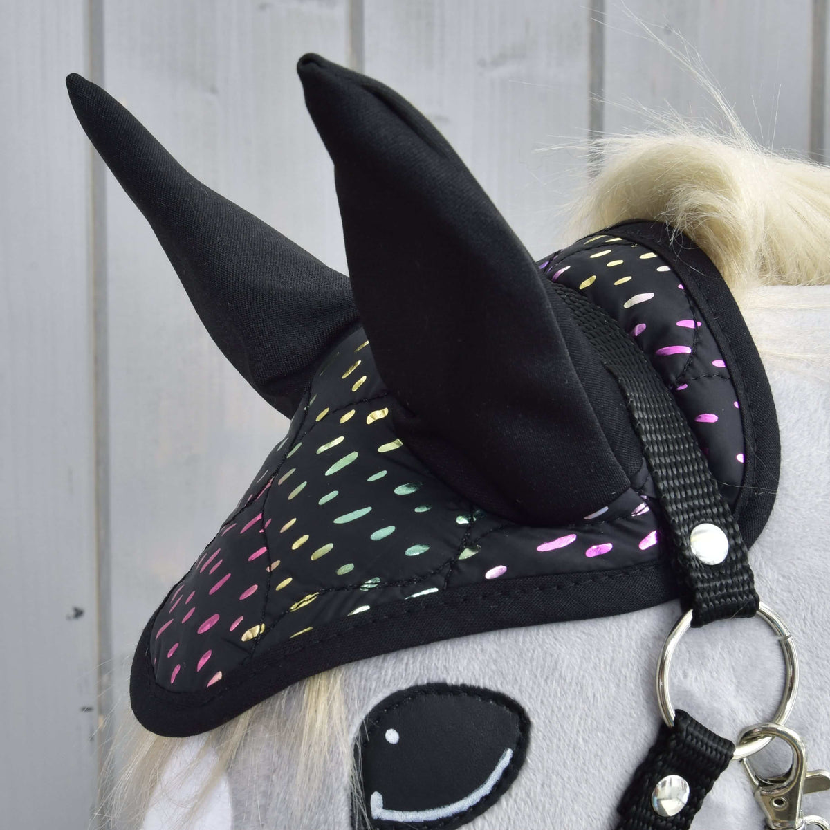Close-up of the Hobby Horse LarDen "Set Dreamy" hobby horse featuring a white body, black ear bonnet with colorful reflective dots, smiling felt eye, and bridle, set against a light wood-paneled background.