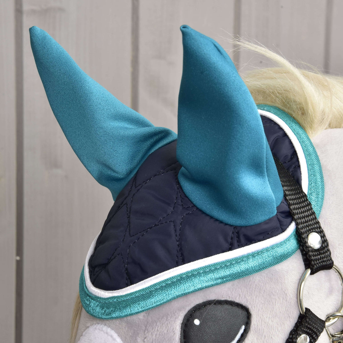 Close-up of Hobby Horse LarDen's Set Supreme Turquoise: a plush toy horse sports a dark blue quilted eye mask with teal pointed ear covers and trim—a stylish ear bonnet set—for fun and protection against a light wood background.