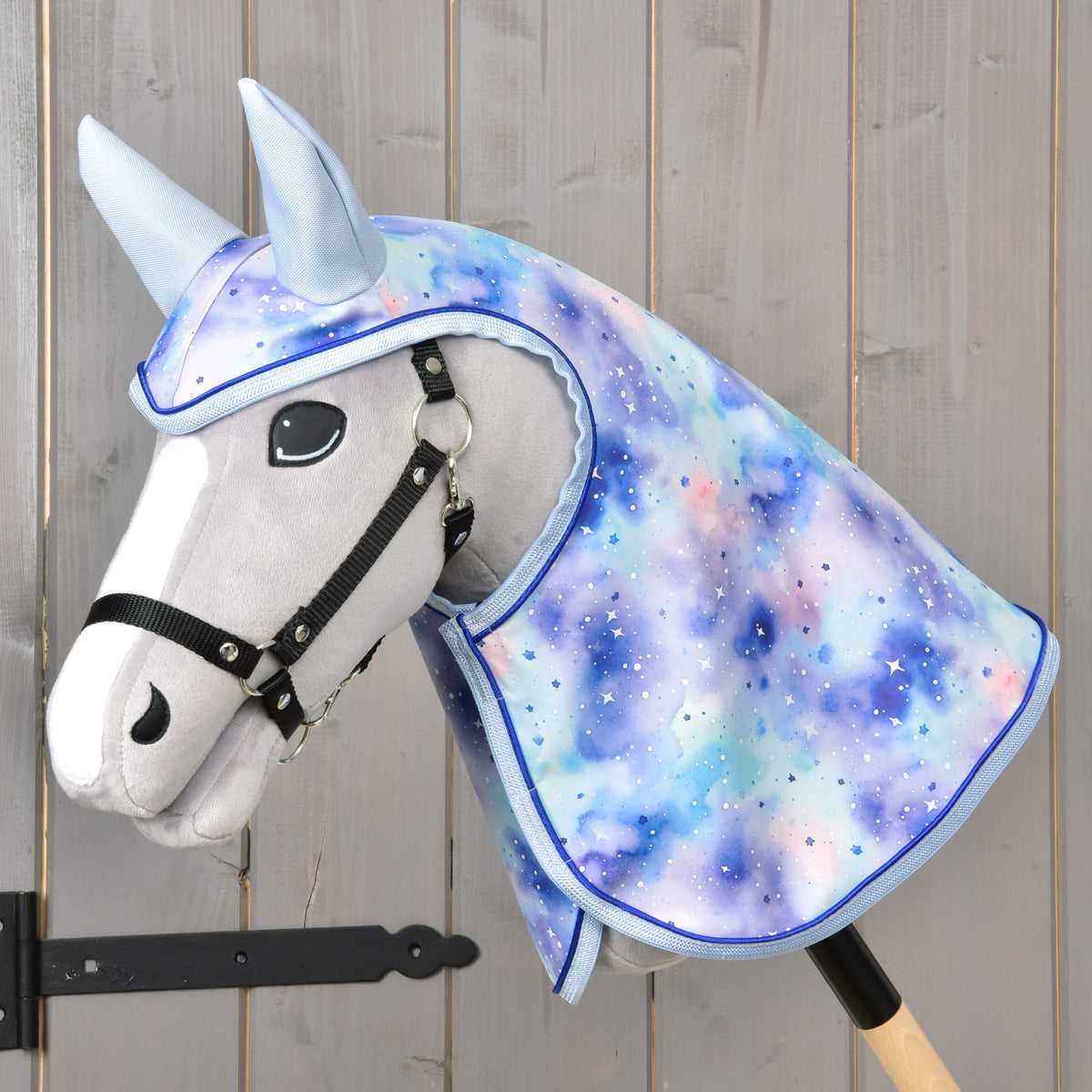 The Hobby Horse LarDen Raincoat Space features a plush hobby horse with a black bridle and a pastel, galaxy-patterned mane and hood in blue, purple, and pink with white speckles. Includes a raincoat for play in any weather. Mounted on a wooden stick.