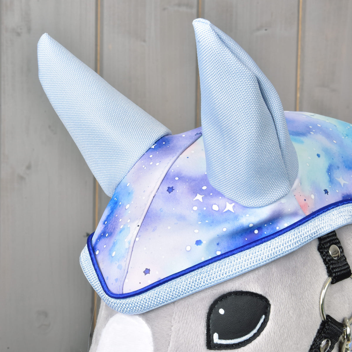 Close-up of Hobby Horse LarDen's Raincoat Space: a stuffed animal wears a hat with tall, light blue horns and a blue-purple galaxy pattern, set against wood—an ideal display for this all-weather raincoat accessory.
