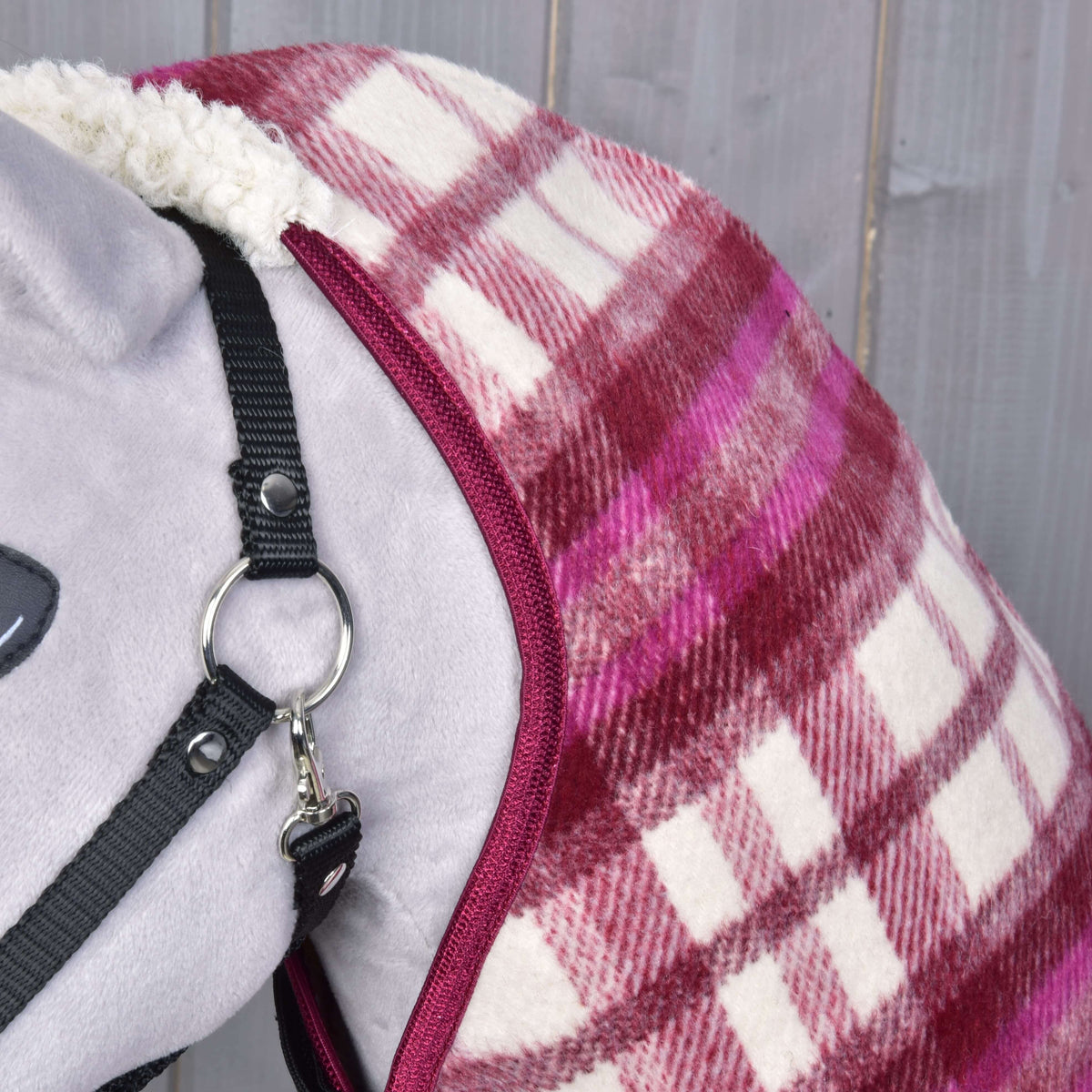 A close-up of a gray horse in front of wooden boards, wearing the Hobby Horse LarDen Stable Blanket in red, pink, and white plaid with white fleece lining and a halter.