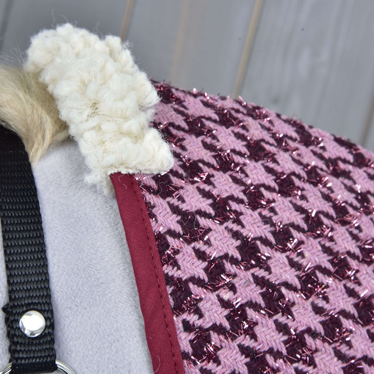 Close-up of the Stable Blanket Winter Magic by Hobby Horse LarDen: a textured pink and purple houndstooth design with sparkly threads, red trim, fluffy white collar, PES material draped on gray with a black strap and metal ring.