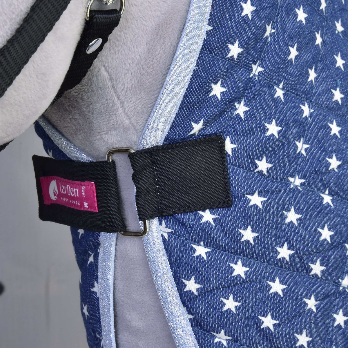 Close-up of the blue quilted Stable Blanket Star by Hobby Horse LarDen, featuring white stars, a black strap with silver buckle, and a pink Izefan label, worn by a gray animal as part of its harness or vest.