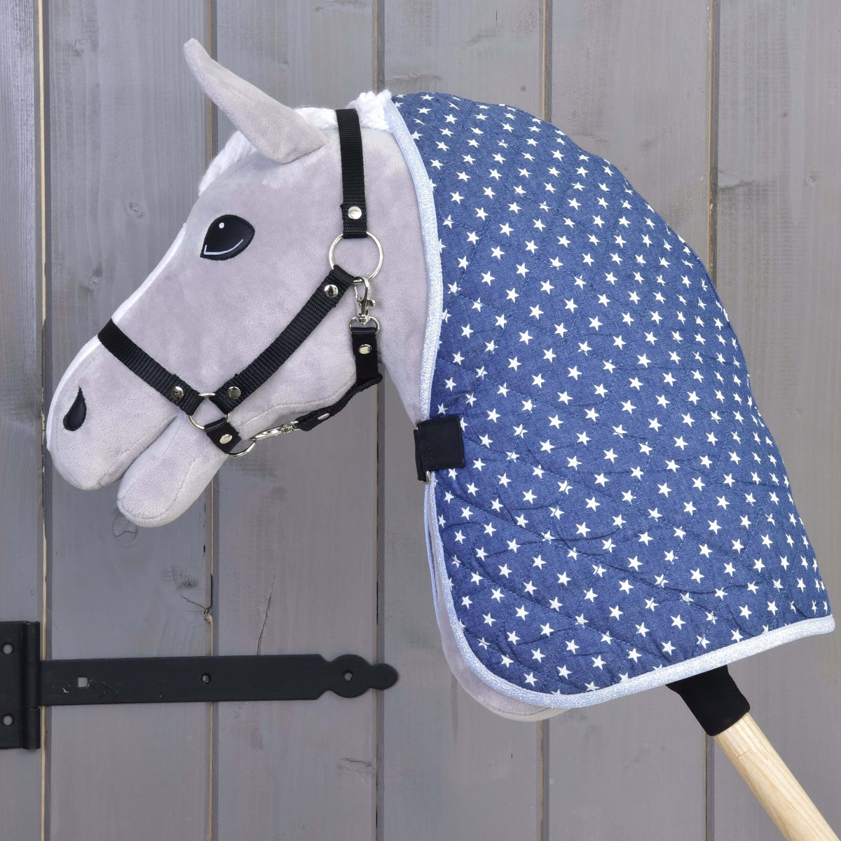 A gray hobby horse toy by Hobby Horse LarDen, featuring a black bridle and the Stable Blanket Star, is mounted on a wooden stick and set against a gray wooden door.
