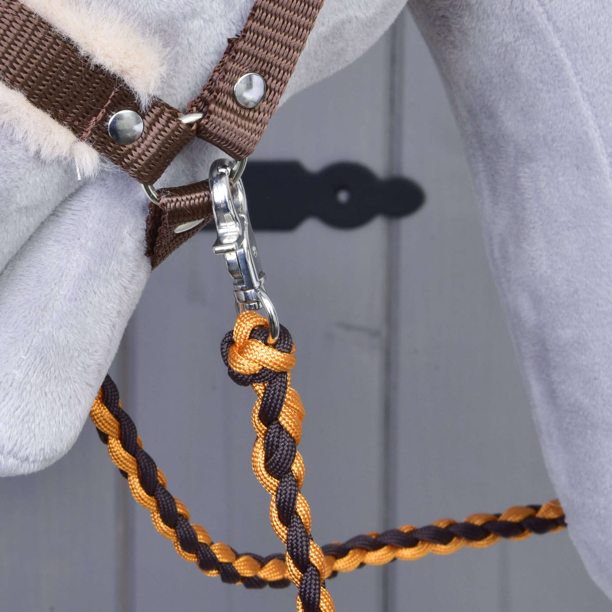 Close-up of a horse’s head wearing the Set Perfect Combination Brown by Hobby Horse LarDen, featuring a fur-padded halter and orange-brown braided lead rope with silver clip. Blurred gray door with black keyhole plate in background—ideal for hobby horse fans.