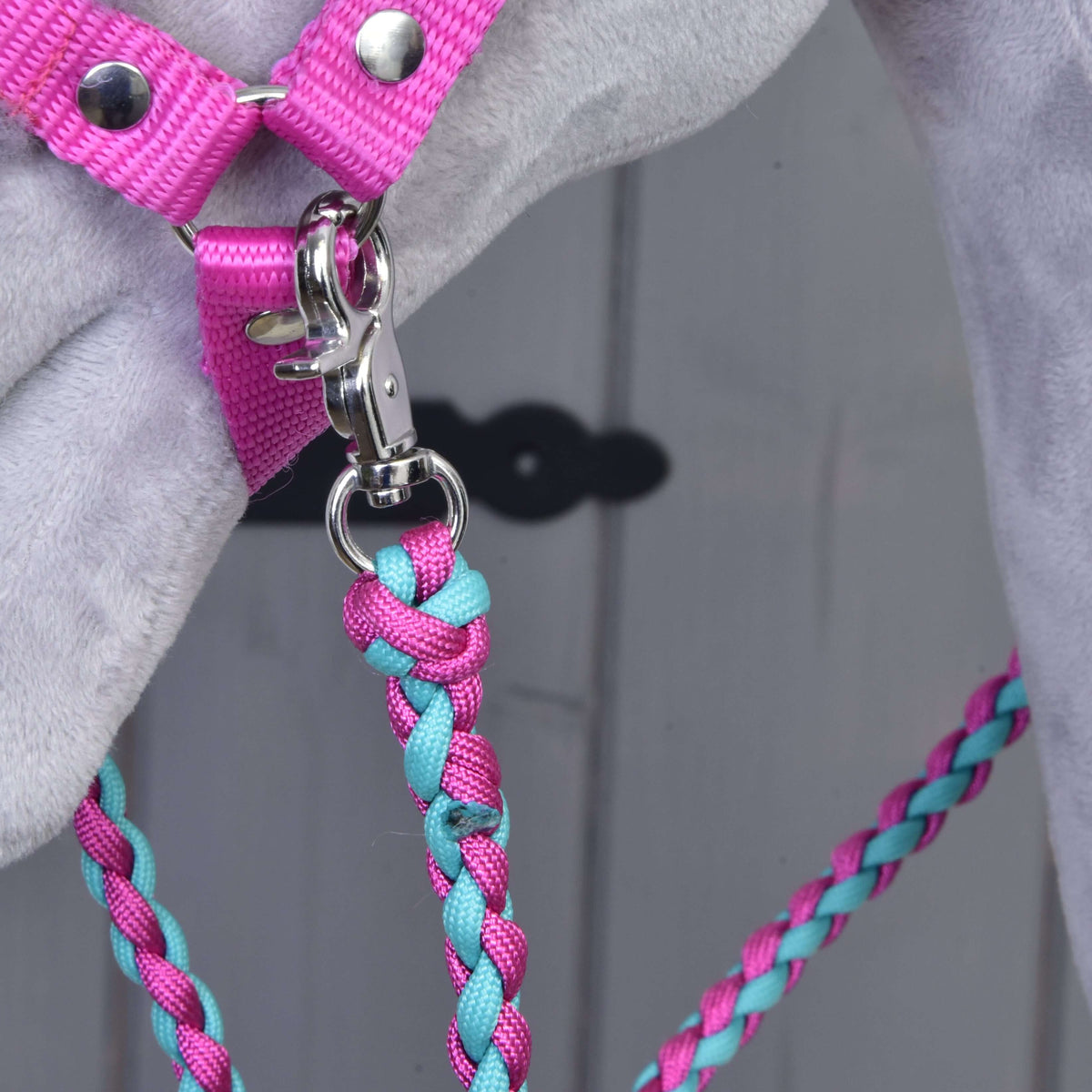 A close-up of the Hobby Horse LarDen Set Perfect Combination Pink—a pink harness and a pink-teal braided lead rope, clipped to a ring on a grey surface—ideal for fans of stylish accessories. A blurred light wood background is visible.