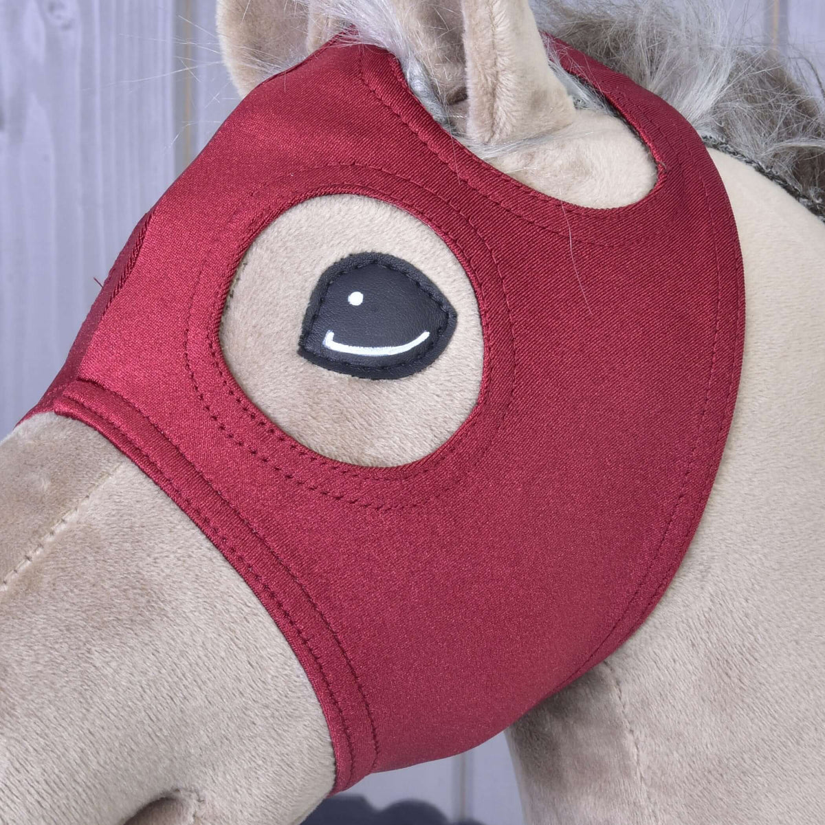 A close-up of a beige hobby horse by Hobby Horse LarDen, featuring a red velvet Horse Mask without Ears that covers its head, with stitched eyes and a fluffy mane visible.