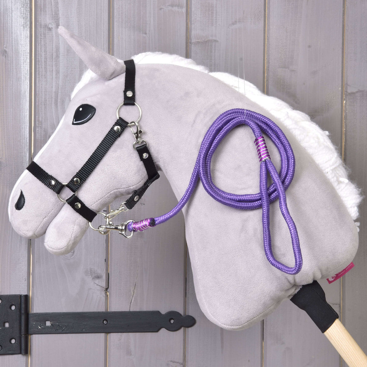The Hobby Horse LarDen "Hobby Horse - rope stripe Viola" features a plush hobby horse with a white mane, black bridle, purple rope leash, and wooden stick, displayed against a light wooden wall.