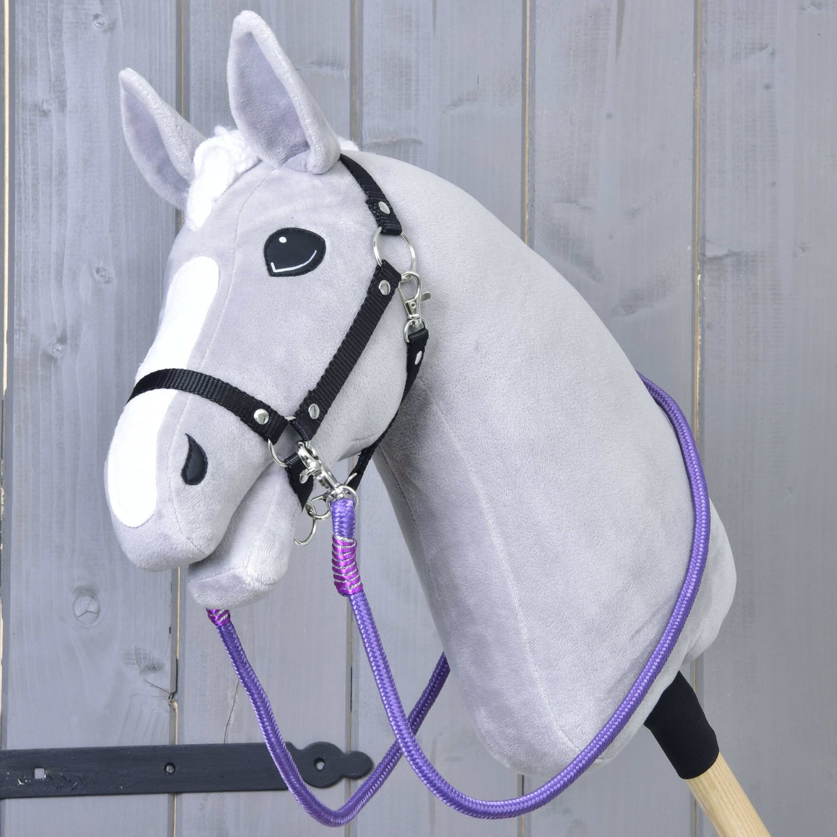 The Hobby Horse LarDen "Reins Viola" is a gray hobby horse with a white blaze, black bridle, and vibrant handmade reins, featuring a plush head on a sturdy stick and displayed against a light wooden fence.