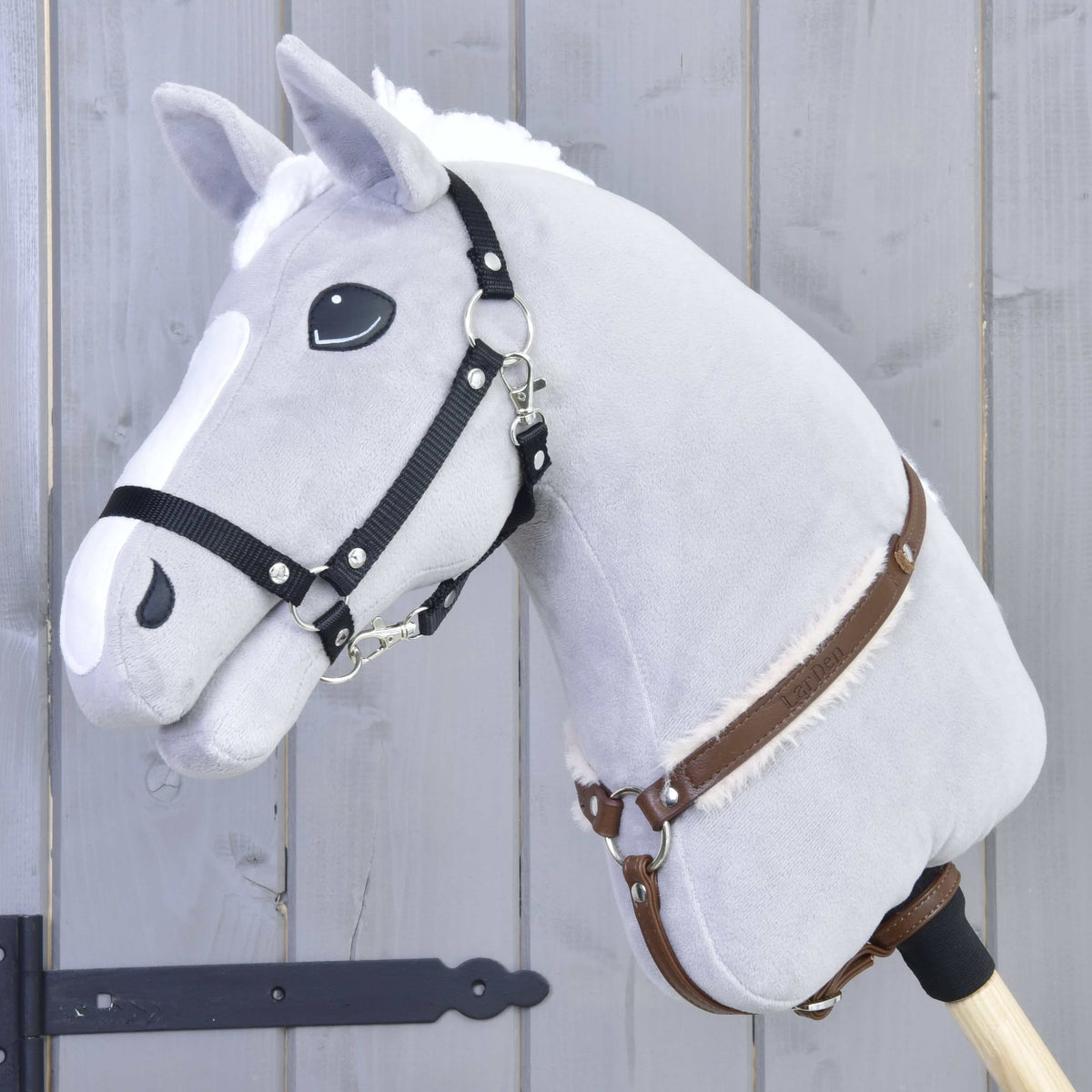 The Hobby Horse LarDen "Breastgirth with fur Walnut" is displayed on a plush gray hobby horse with a white mane, black bridle, leather reins, and wooden stick, set against a gray wooden background.