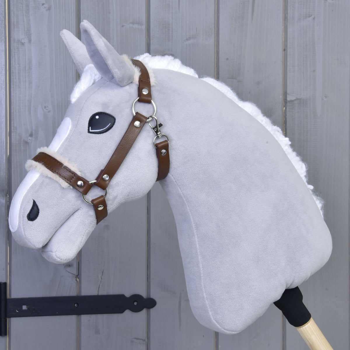 The Hobby Horse LarDen Halter with fur - leatherette Walnut is a gray plush hobby horse with a white mane, brown leatherette bridle, and large black eyes on a wooden stick, shown leaning against a gray wooden wall.
