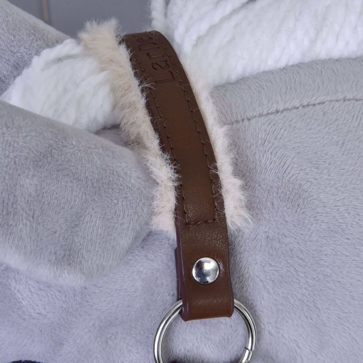 Close-up of the Hobby Horse LarDen Halter with fur - leatherette Walnut, featuring plush grey and white fabric joined by a fluffy seam. A brown leatherette strap, fastened with a silver button and ring, lies across the junction.