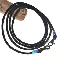 The Hobby Horse LarDen Western Reins Montana features a black braided cord with snap hooks at both ends, accented by blue, green, and pink bands—ideal for training or western riding. Coiled in a circle on white background with wood at the top left.