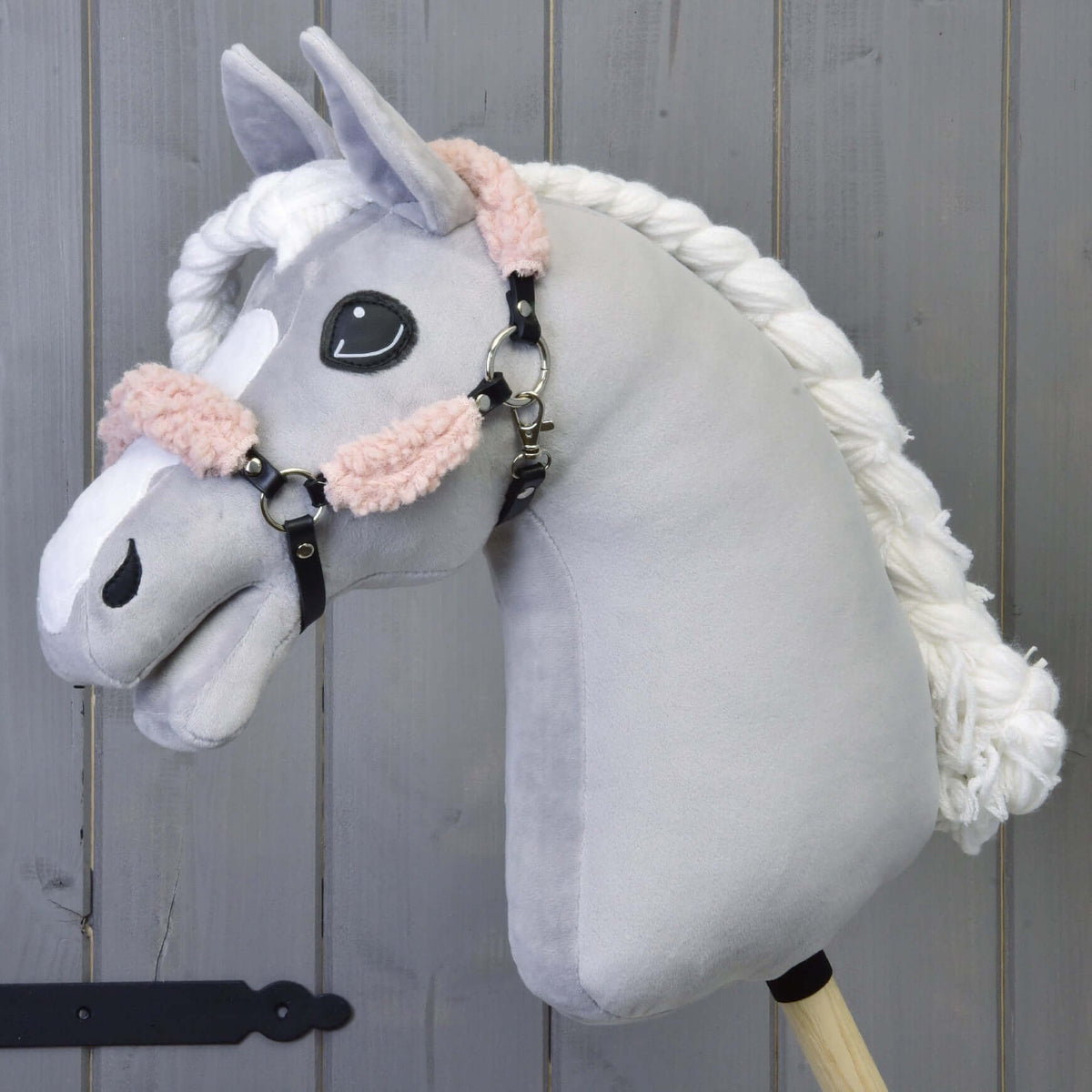 The Hobby Horse LarDen Black Halter with Old Rose Fur Complete – leatherette is a plush hobby horse with a light gray head, white braided mane, and black halter, mounted on a wooden stick against a wooden panel background.