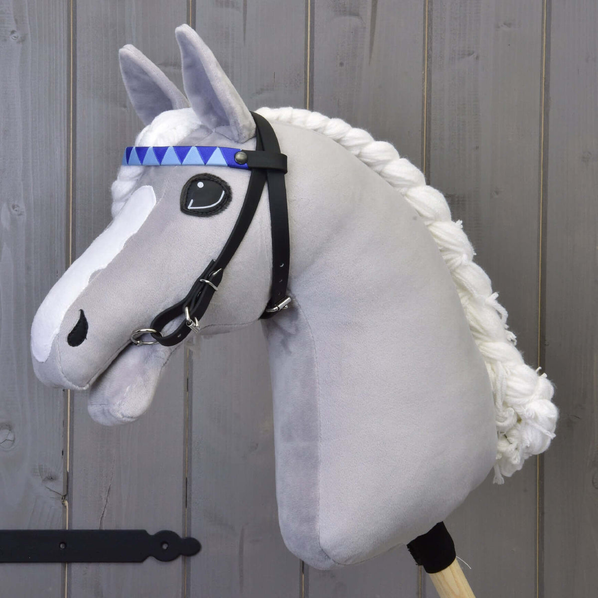 The Hobby Horse LarDen Bridle with 2 Headbands Blue (size M) features a gray plush horse with a braided white mane, black bridle, blue patterned headband, detachable reins, and is mounted on a wooden stick against a wooden wall.