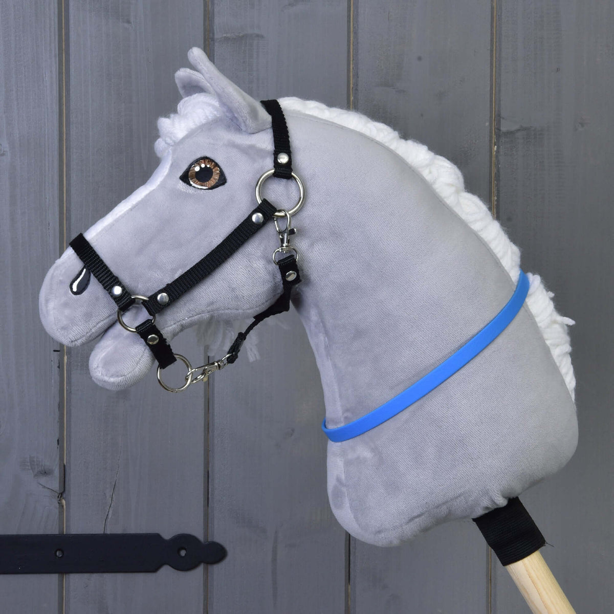 The Hobby Horse LarDen "Hobby Horse cordeo blue (size S)" features a gray plush body, white mane, black bridle, and blue-violet ribbon on a wooden stick, displayed against a gray wooden wall.