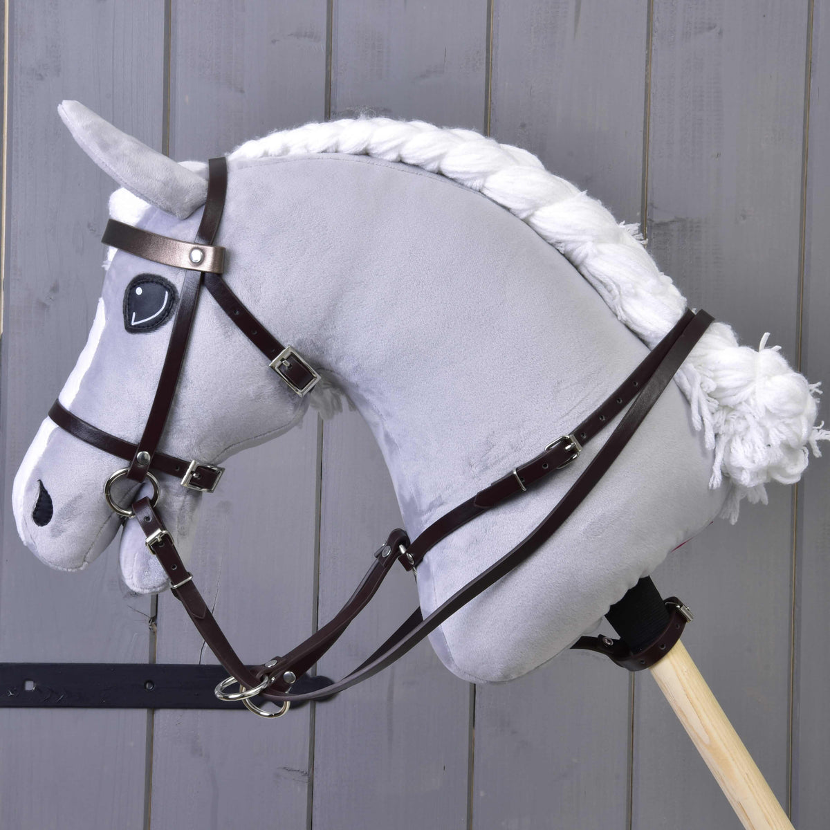 The Hobby Horse LarDen Martingale brown, a gray plush hobby horse with white yarn mane and tail, black bridle, and leatherette martingale on a wooden stick, is shown against a gray wooden plank background.